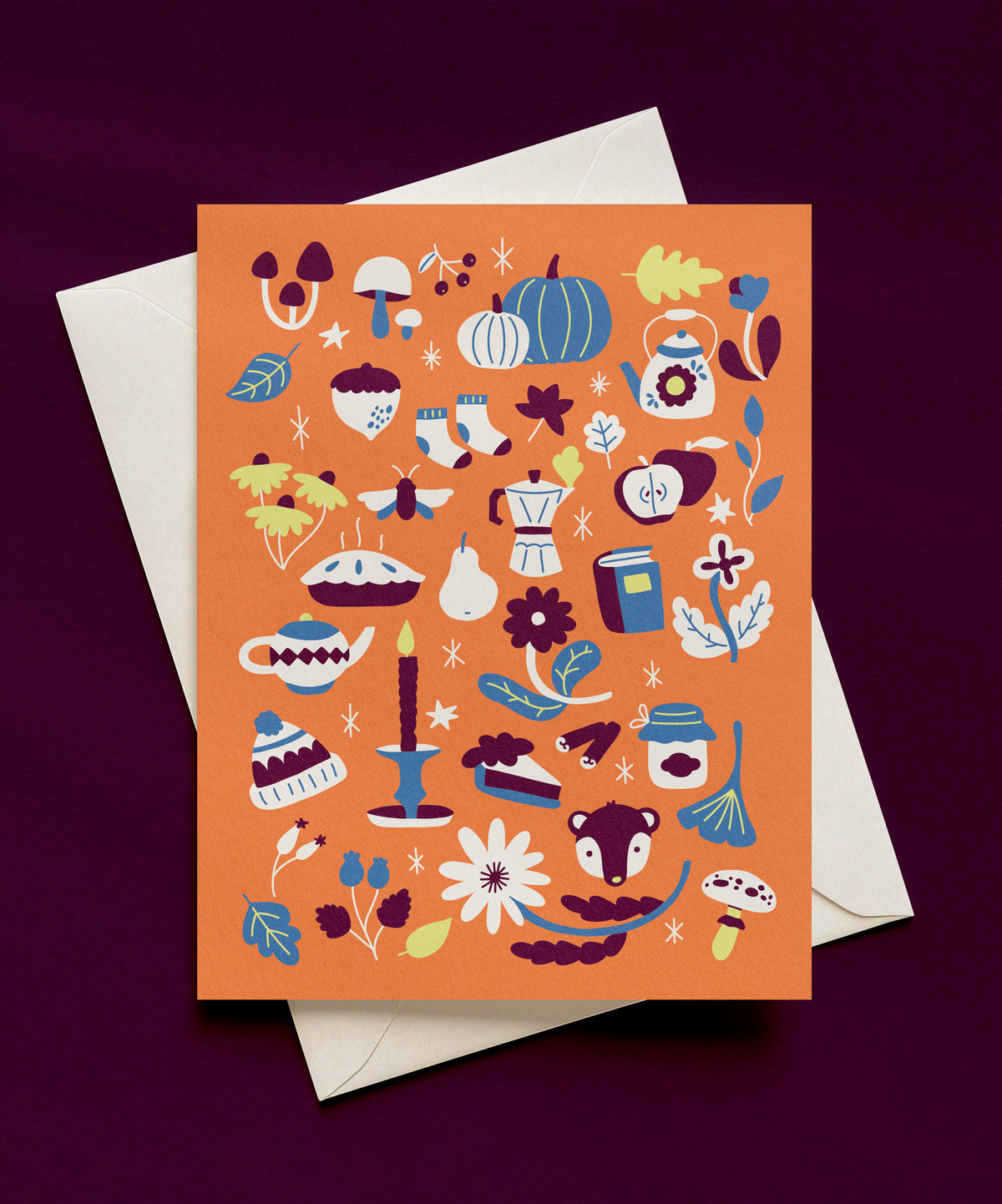 Orange greeting card covered in autumn illustrations including pumpkins, mushrooms, leaves, apples, pie, a teapot, a candle, and a knit hat, shown with an envelope