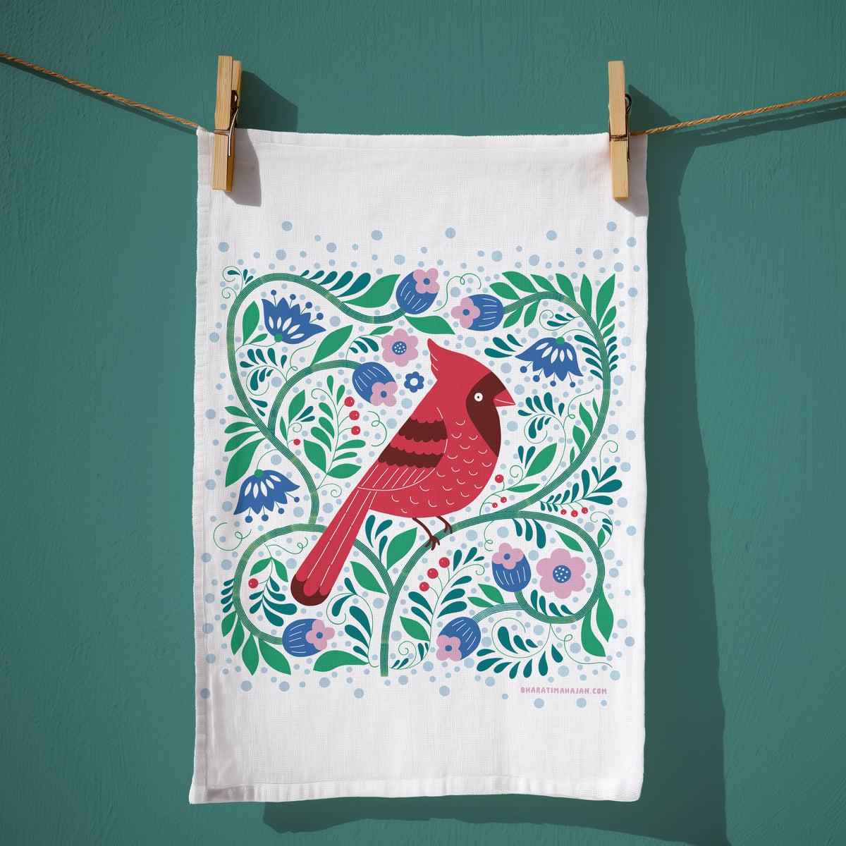 A white kitchen towel with a colorful folk-art style illustration of a red cardinal bird and green floral vines, hanging from a rope with wooden clips against a teal wall.