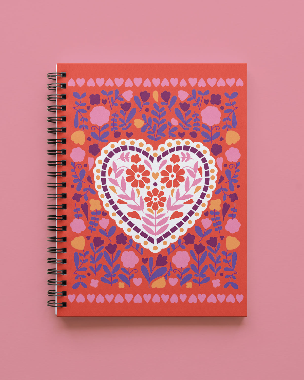 Wire bound notebook with a coral-red cover showing a white scalloped heart filled with orange and pink flowers, surrounded by purple and pink florals and a border of small hearts, shown on a pink background