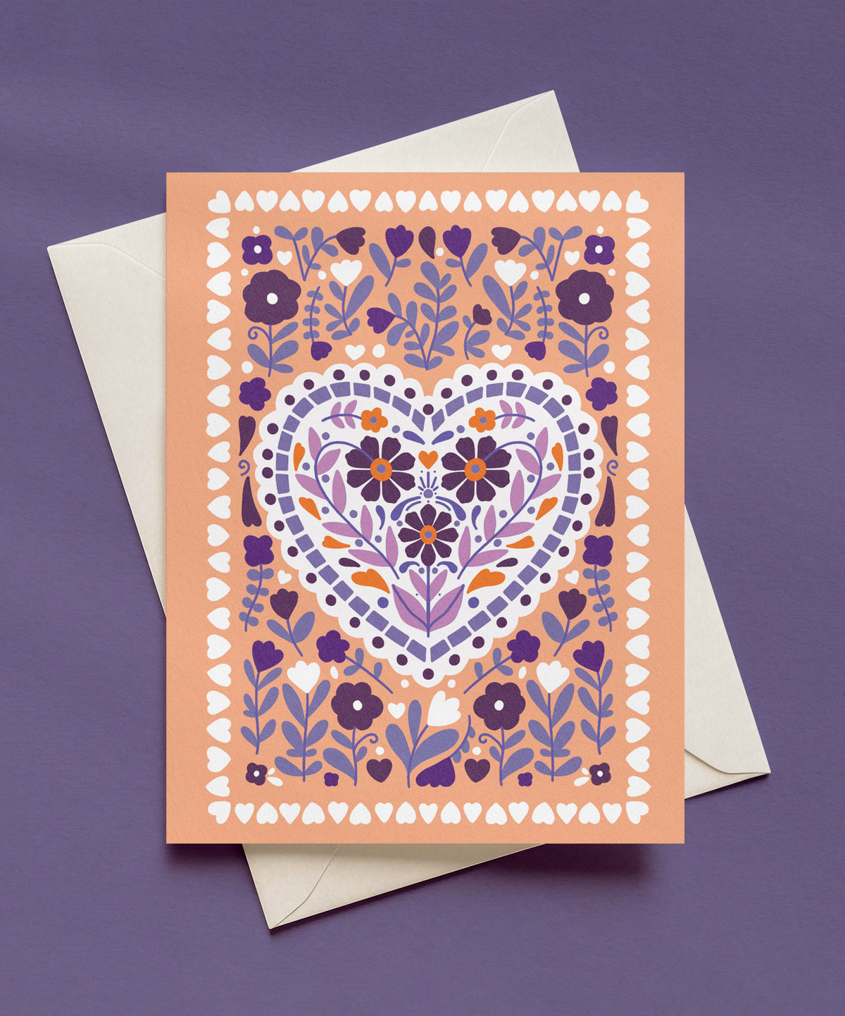 Peach greeting card with a white scalloped heart filled with plum and lavender flowers, surrounded by purple florals and a border of small white hearts, shown with a cream envelope