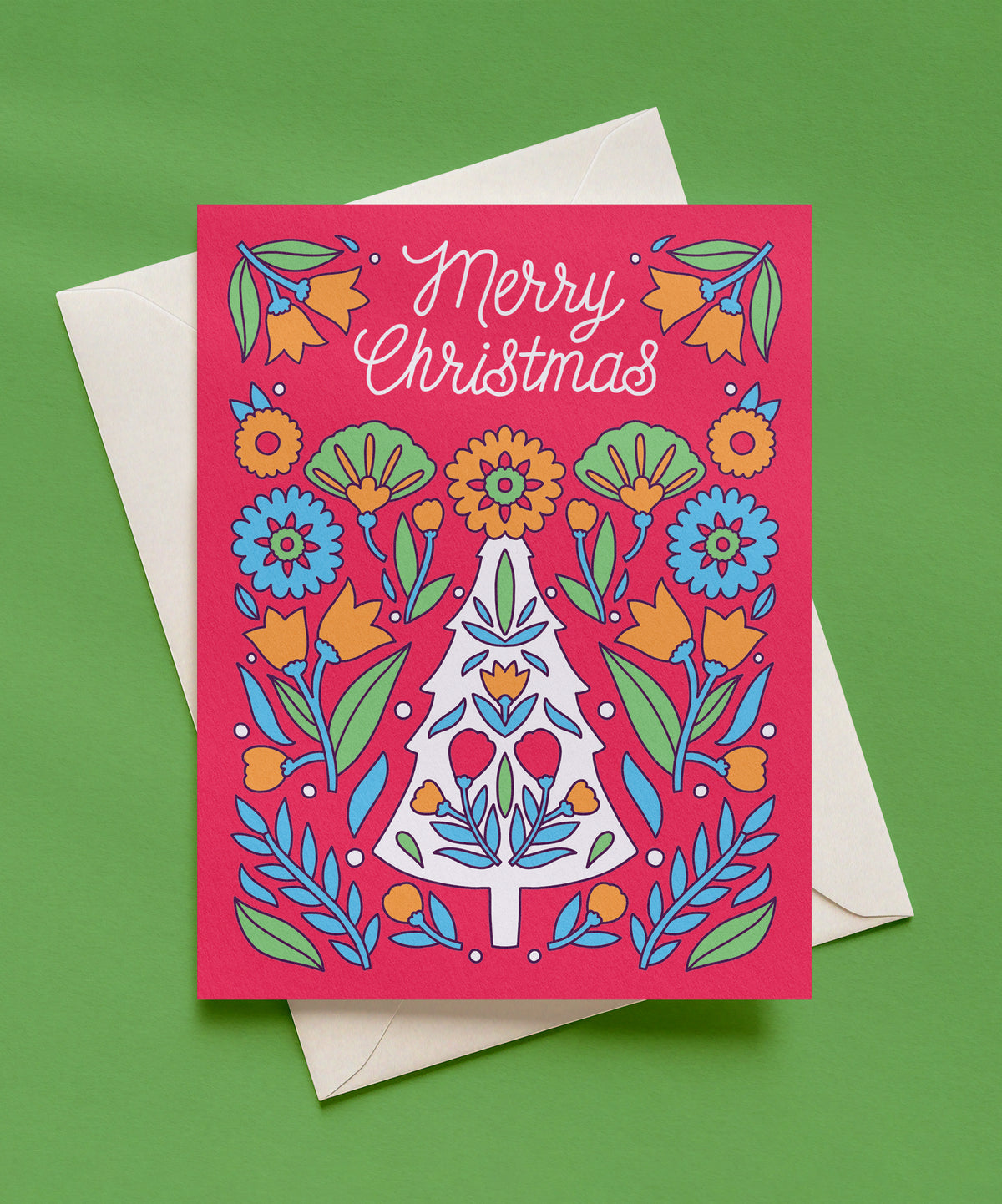 Pink Merry Christmas greeting card with white script lettering, a white Christmas tree filled with folk-style flowers, and orange, blue, and green florals, shown with a cream envelope