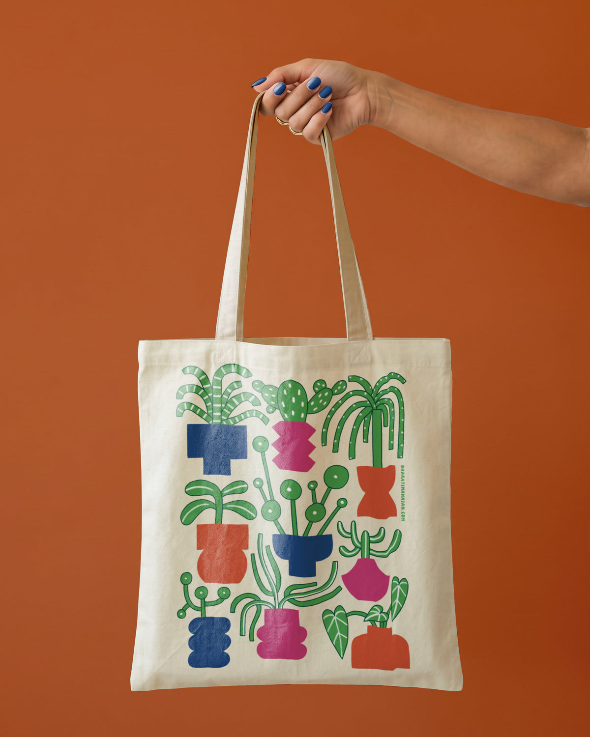 Natural canvas tote bag with a colorful illustration of potted houseplants and cacti in blue, pink, and orange pots, held by one hand against a terracotta background