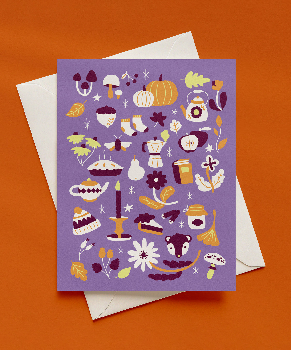 Purple greeting card covered in autumn illustrations including pumpkins, mushrooms, leaves, apples, pie, a teapot, a candle, and a knit hat, shown with an envelope