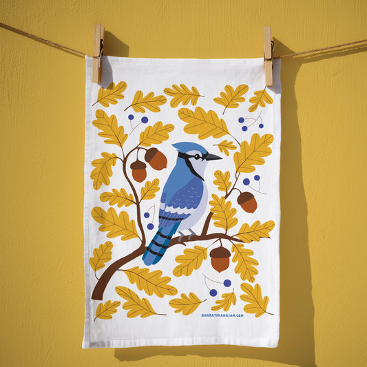 Cotton kitchen towel with a blue jay perched on an oak branch among golden leaves, acorns, and blue berries, hanging from wooden clothespins