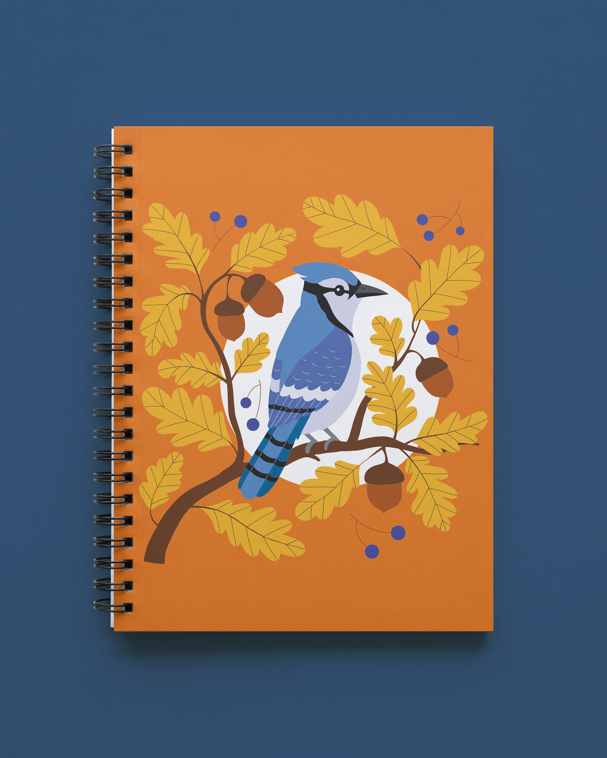 wire-bound notebook with an orange cover showing a blue jay perched on an oak branch among golden leaves, acorns, and blue berries, shown on a deep blue background