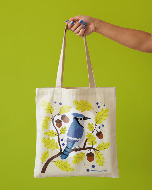 Natural tote bag with a blue jay perched on an oak branch among green leaves, acorns, and blue berries, held by one hand against a lime green background