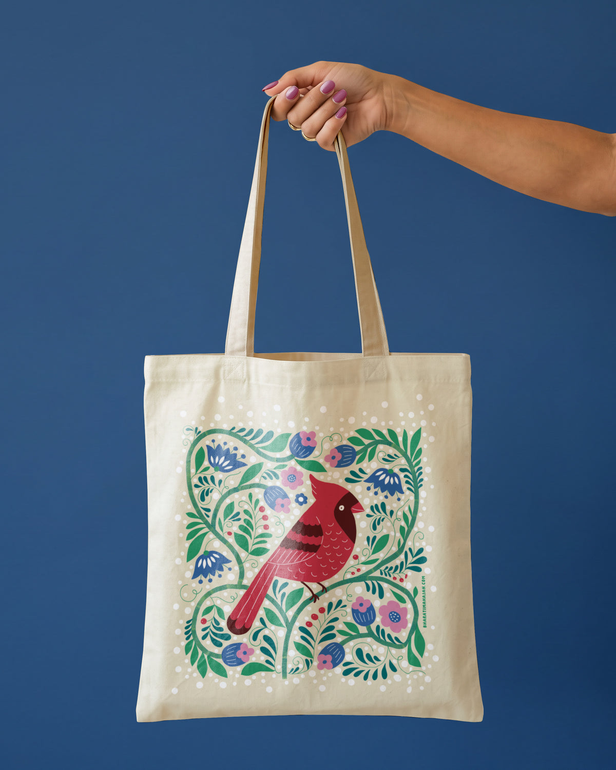 Natural tote bag with a red cardinal surrounded by blue and pink flowers, green vines, and berries, held by one hand against a deep blue background