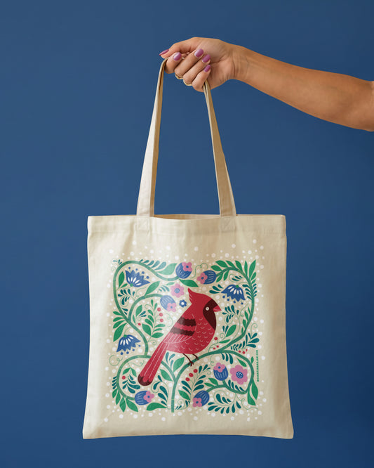 Natural tote bag with a red cardinal surrounded by blue and pink flowers, green vines, and berries, held by one hand against a deep blue background