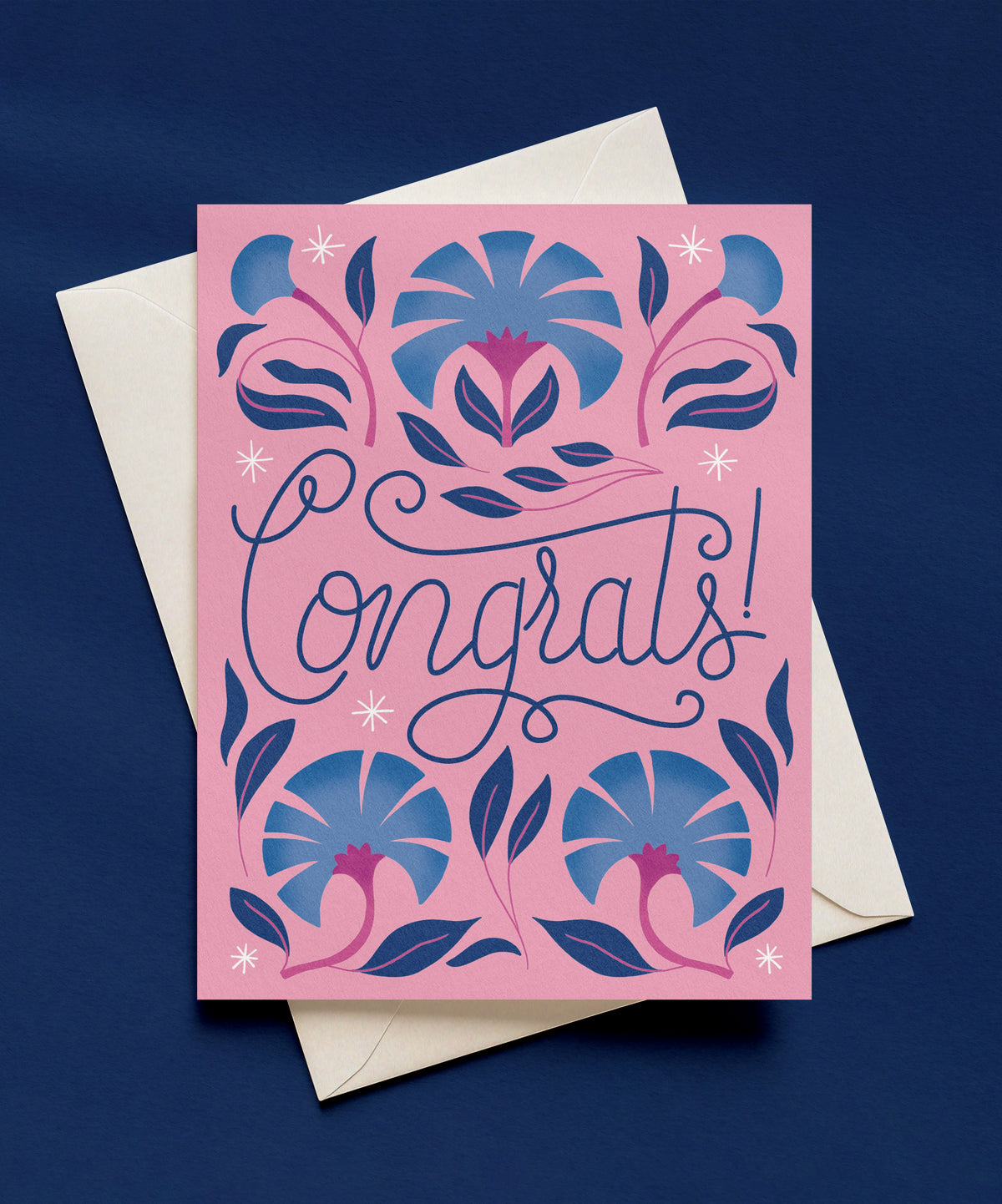 Congrats greeting card with navy script lettering on a pink background, surrounded by blue and magenta flowers and white sparkles, shown with a cream envelope