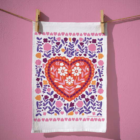 Cotton kitchen towel with a red heart filled with white and pink flowers, surrounded by purple, pink, and orange florals and a border of small hearts, hanging from wooden clothespins