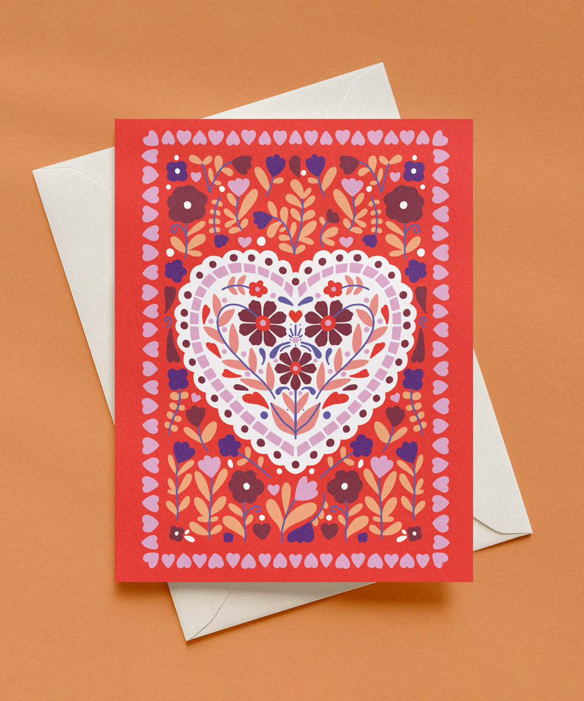 Red greeting card with a white scalloped heart filled with maroon and coral flowers, surrounded by purple and peach florals and a border of small pink hearts, shown with a cream envelope