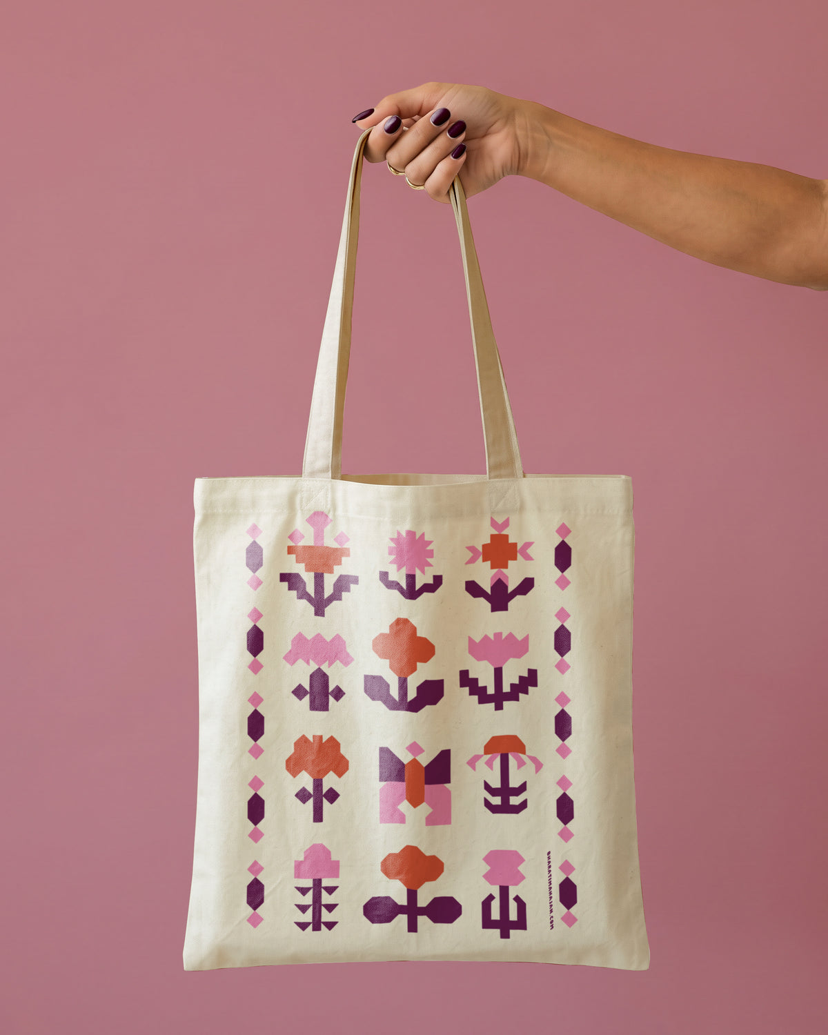 Natural tote bag with a geometric flower pattern in pink, coral, and plum, held by one hand against a dusty rose background