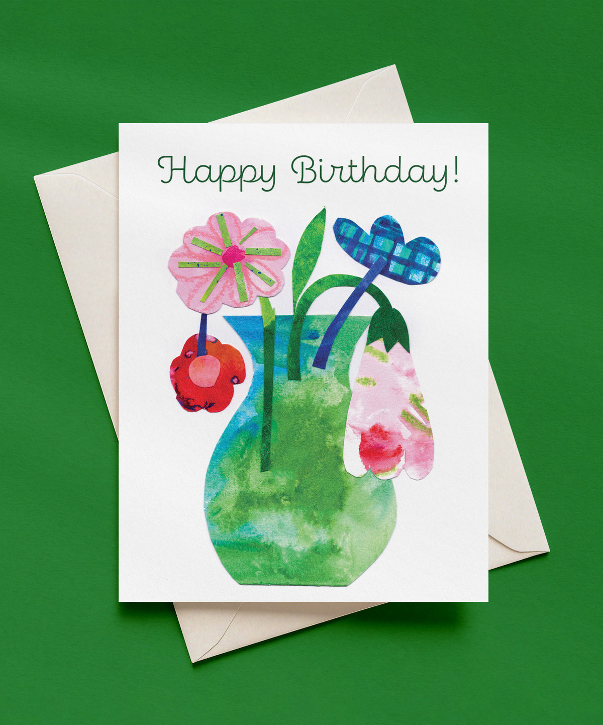Happy Birthday greeting card with green script lettering above a collage-style green and blue vase holding pink, red, and plaid blue flowers on a white background, shown with a cream envelope