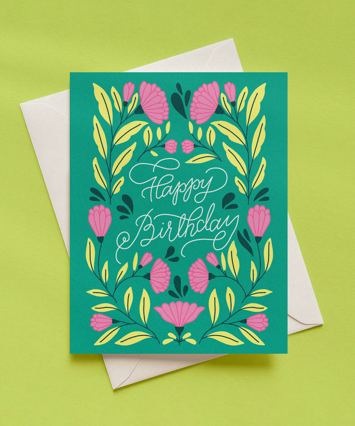 Emerald green Happy Birthday greeting card with white script lettering, surrounded by pink flowers and yellow-green leaves, shown with a cream envelope