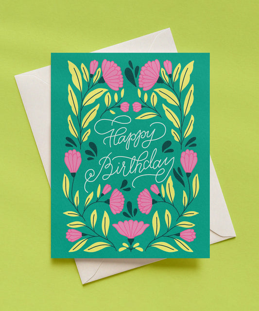 Emerald green Happy Birthday greeting card with white script lettering, surrounded by pink flowers and yellow-green leaves, shown with a cream envelope