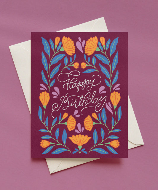 Happy Birthday greeting card with white script lettering on a plum background, surrounded by orange flowers and blue leaves, shown with a cream envelope