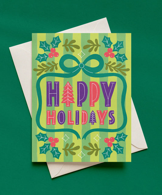 Happy Holidays greeting card with purple and pink lettering, a teal bow, holly leaves, and red berries on a green striped background, shown with a cream envelope