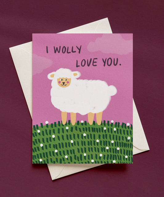 I Wolly Love You greeting card with a fluffy white sheep on a green flowery hill under a pink sky, shown with a cream envelope