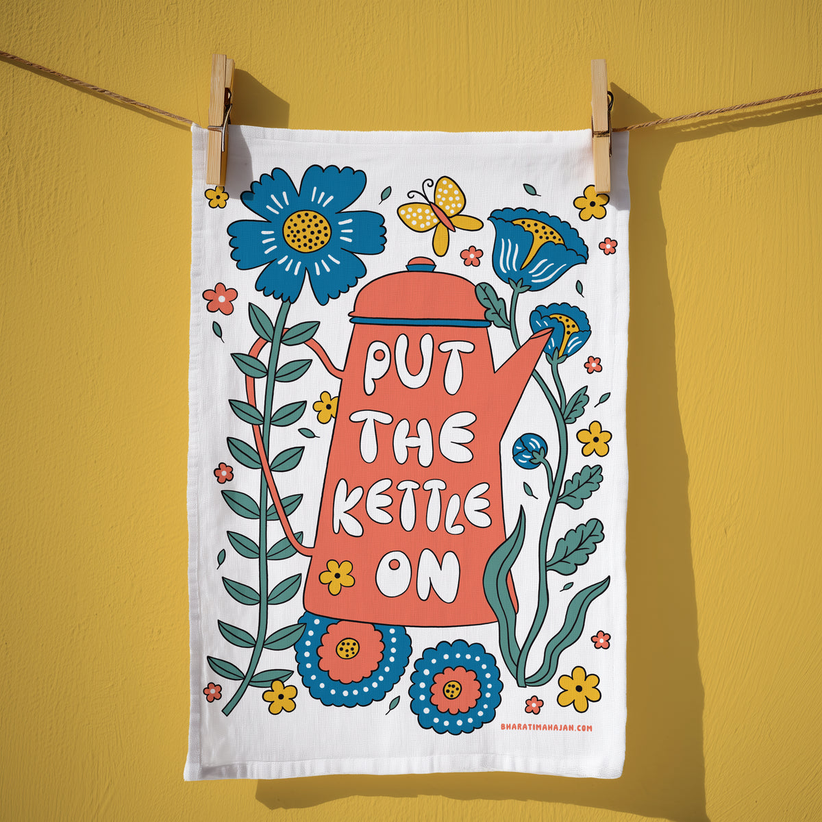 Cotton kitchen towel with a coral kettle and the words Put the Kettle On, surrounded by blue and yellow flowers and a butterfly, hanging from wooden clothespins
