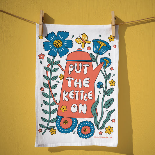 Cotton kitchen towel with a coral kettle and the words Put the Kettle On, surrounded by blue and yellow flowers and a butterfly, hanging from wooden clothespins