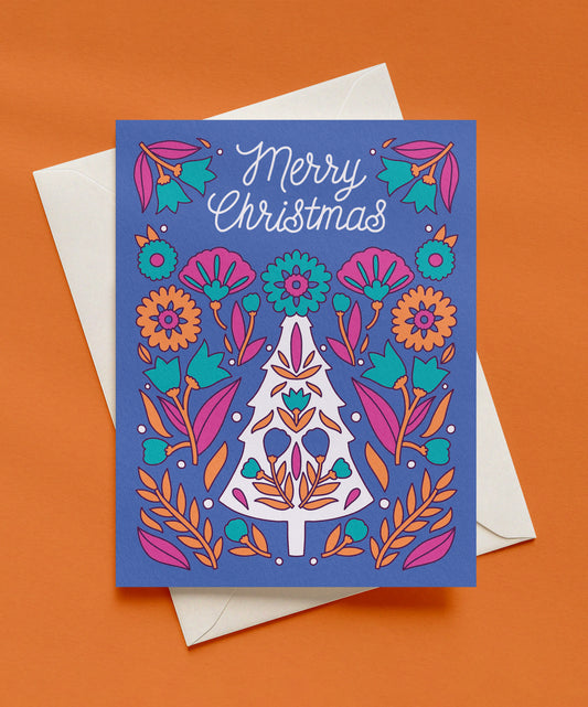 Blue Merry Christmas greeting card with white script lettering, a white Christmas tree filled with folk-style flowers, and magenta, orange, and teal florals, shown with a cream envelope