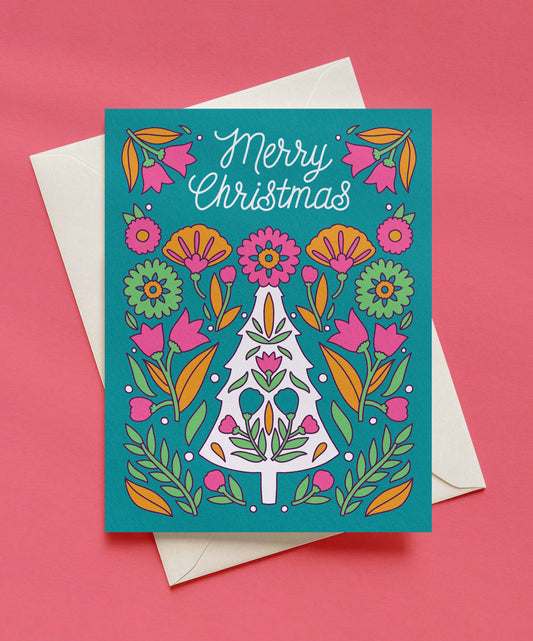 Teal Merry Christmas greeting card with white script lettering, a white Christmas tree filled with folk-style flowers, and pink, orange, and green florals, shown with a cream envelope