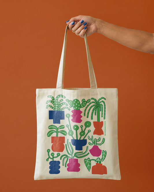 Natural canvas tote bag with a colorful illustration of potted houseplants and cacti in blue, pink, and orange pots, held by one hand against a terracotta background
