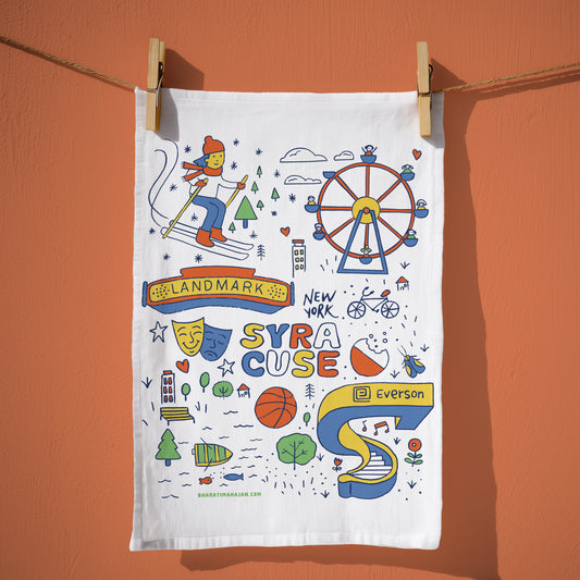 A white fabric tea towel with colorful New York City-themed illustrations hangs from wooden clothespins on a rope against a solid terracotta wall. The towel features drawings of a skier, a Ferris wheel, a theater mask, a basketball, and the text 'SYRACUSE' ,'LANDMARK','New York' and 'Everson'.