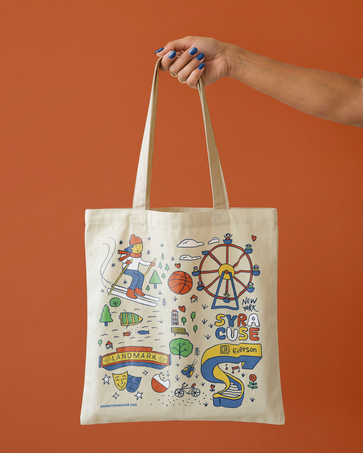 Natural tote bag with a hand-drawn Syracuse, New York design featuring a skier, Ferris wheel, Landmark Theatre sign, theater masks, and Everson Museum, held by one hand against a terracotta background